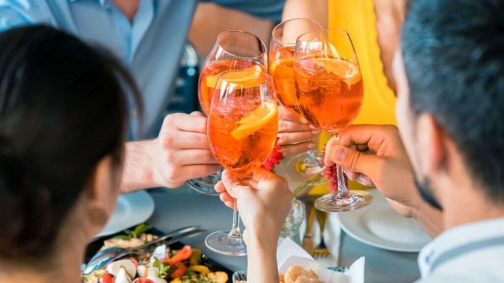 Four people clinking glasses of orange-colored cocktails with orange slices during happy hour at a table set with food.
