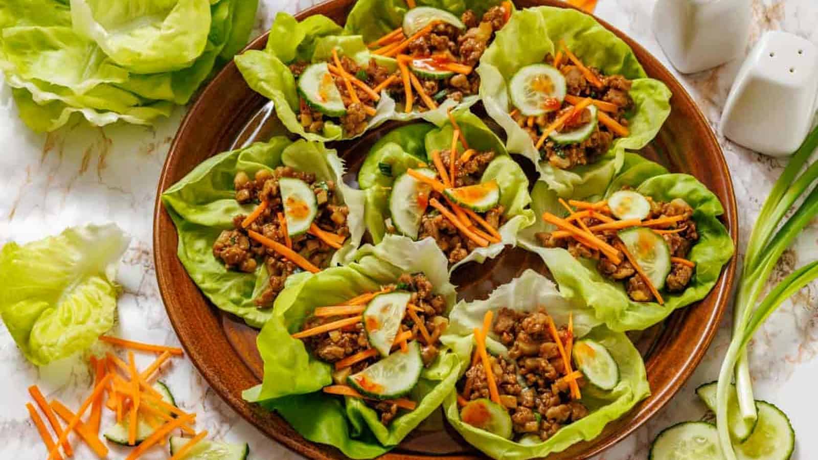 A wooden plate with Chicken lettuce wraps surrounded by extra lettuce leaves and vegetables on a marble surface.
