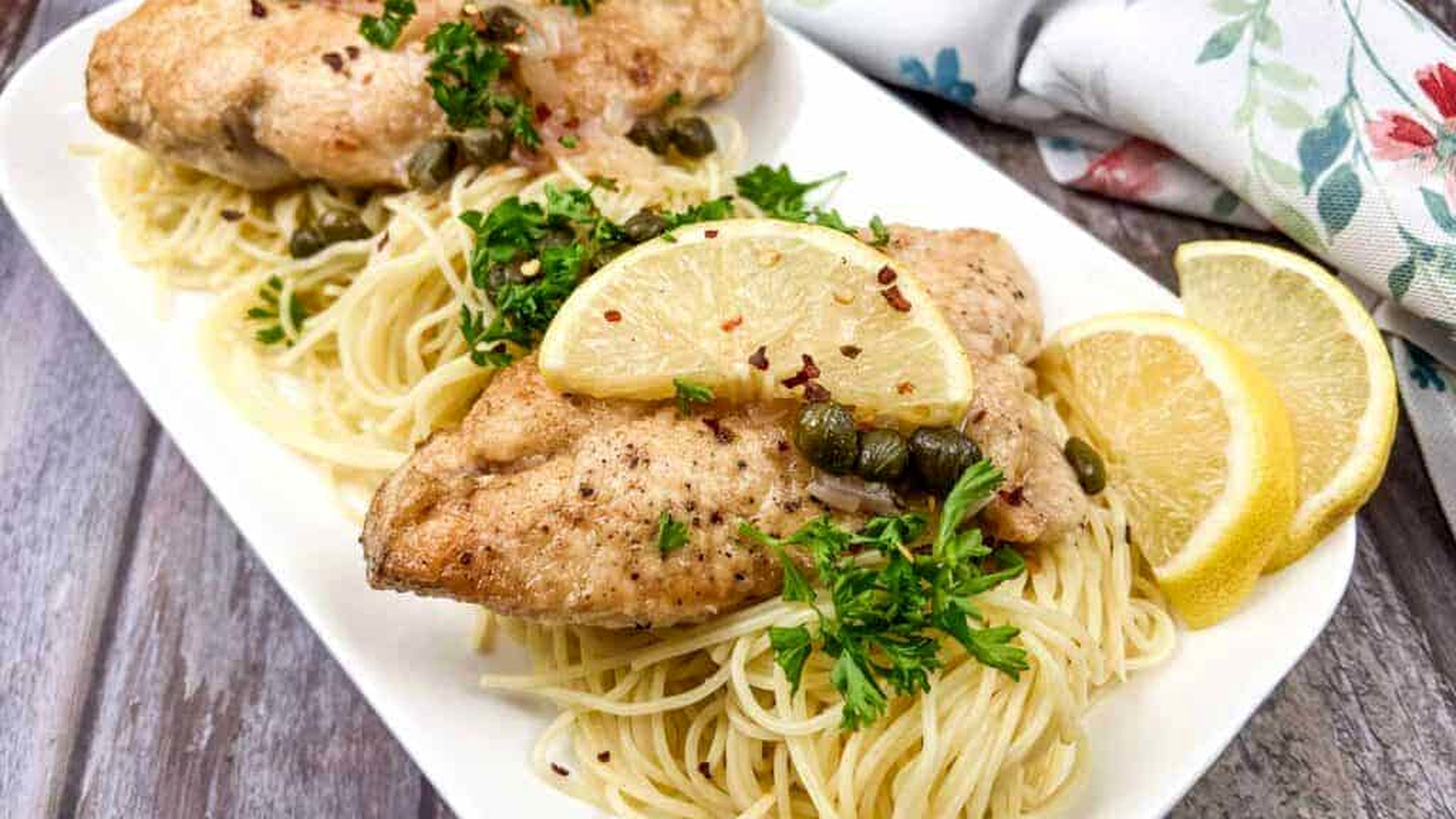 Chicken Piccata served over pasta, garnished with lemon slices, capers, and parsley.