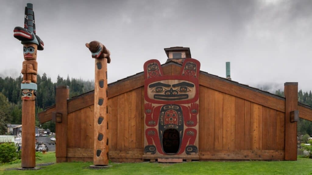 A traditional wooden longhouse with colorful Indigenous carvings and two totem poles on a grassy area, set against a forested, misty background—a scene you might glimpse on a Windstar Alaska cruise.