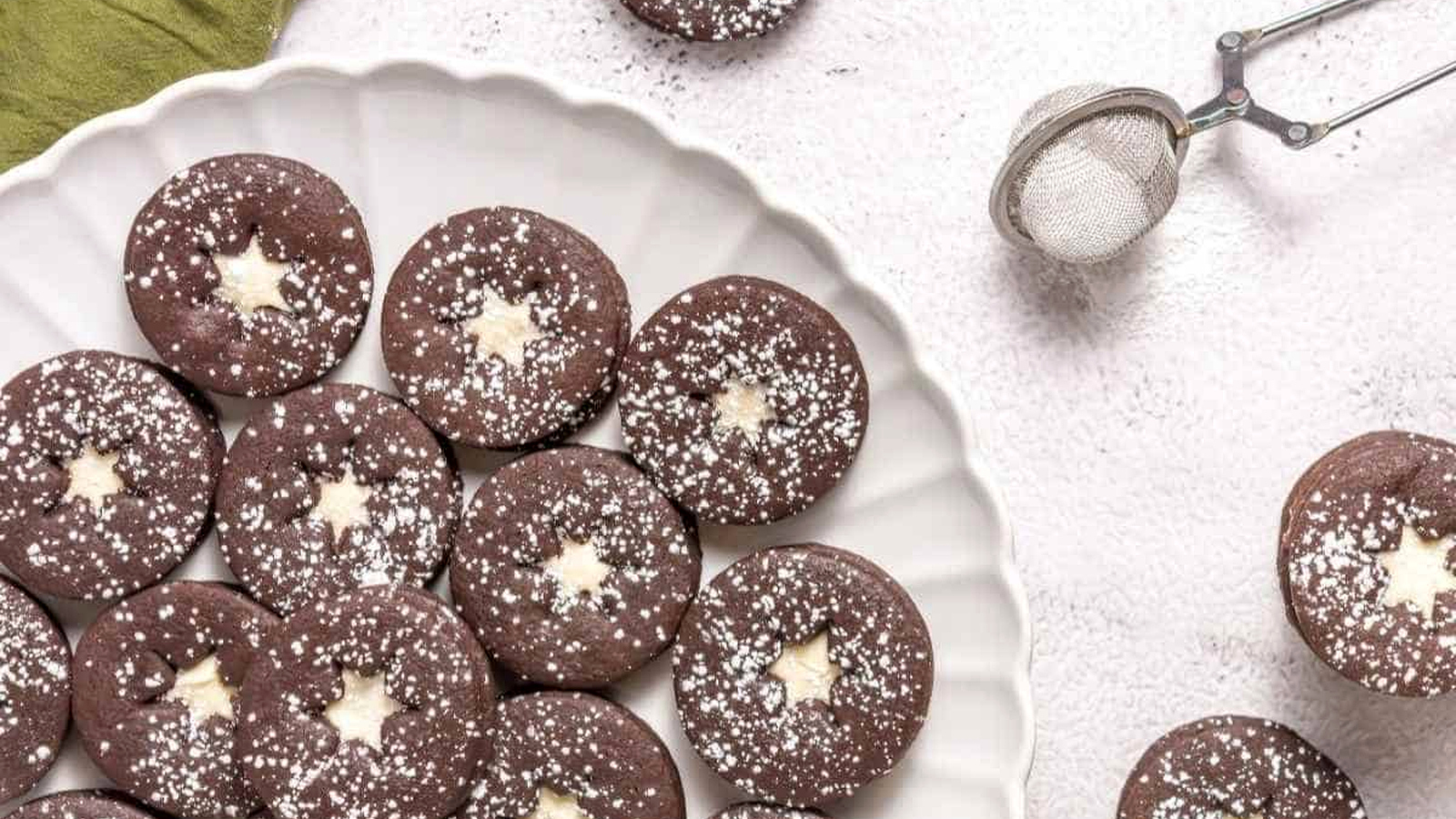 A plate of round chocolate cookies with star-shaped centers, dusted with powdered sugar. A metal sifter with powdered sugar is on the side.