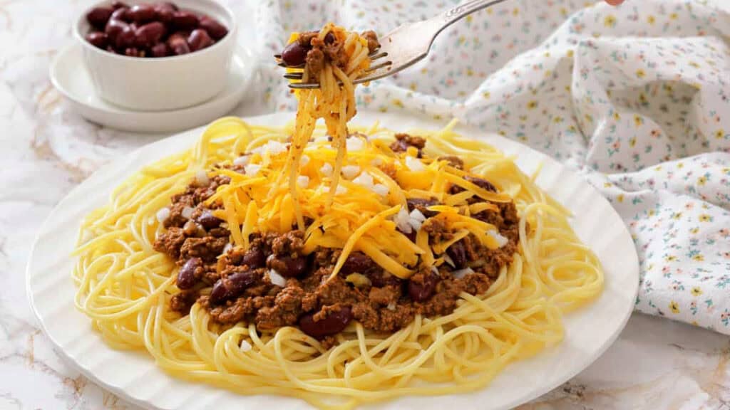 A plate of spaghetti topped with chili, kidney beans, shredded cheddar cheese, and chopped onions. A fork is lifting a portion of the noodles. A bowl of beans is in the background.