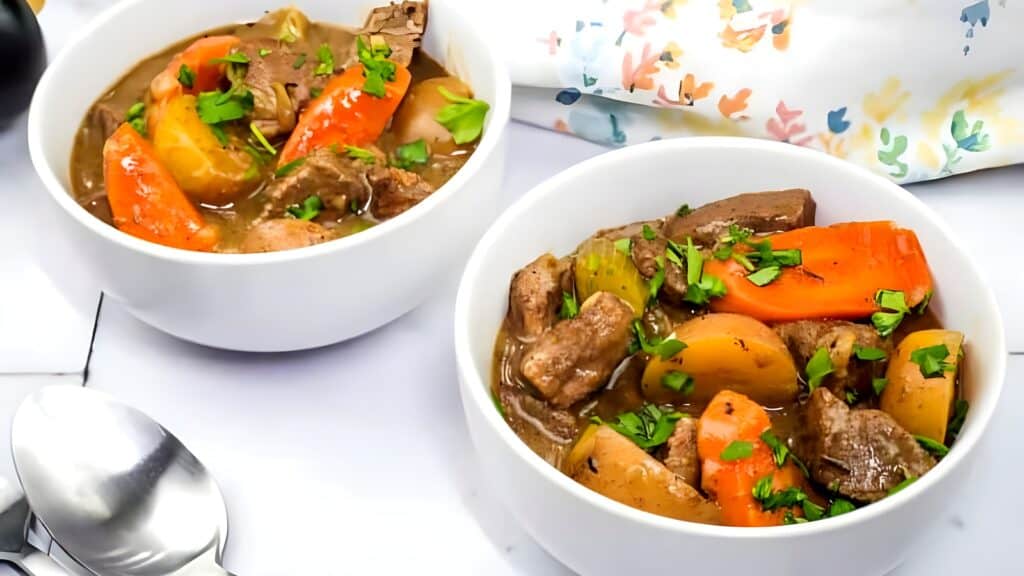 Two white bowls filled with beef stew, containing chunks of beef, carrots, potatoes, and parsley, placed on a white surface with two spoons nearby.