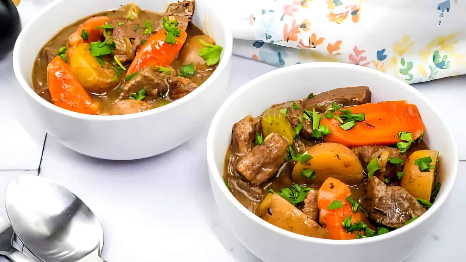 Two bowls of beef stew alongside two silver spoons.