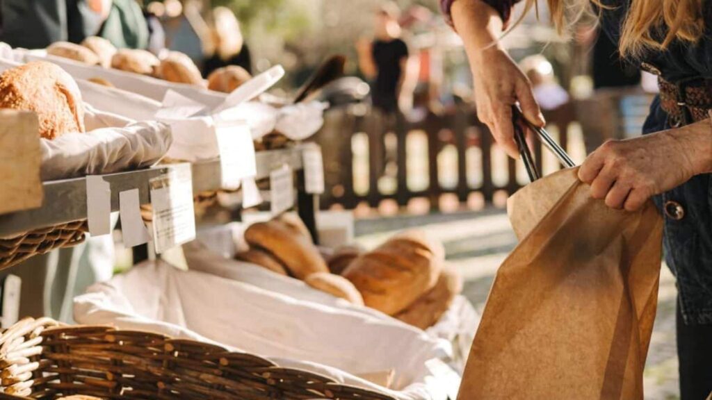 A person places bread into a brown paper bag at an outdoor market stand displaying various loaves of bread.