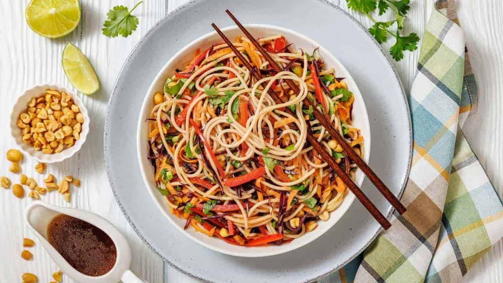 A bowl of Asian noodle salad with vegetables and chopsticks, surrounded by lime, peanuts, sauce, and fresh cilantro on a light wooden table with a plaid napkin.