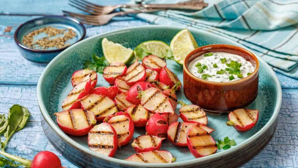 A plate of grilled radishes with grill marks, lime wedges, and a bowl of creamy dip garnished with herbs, set on a blue surface with a napkin and cutlery in the background.