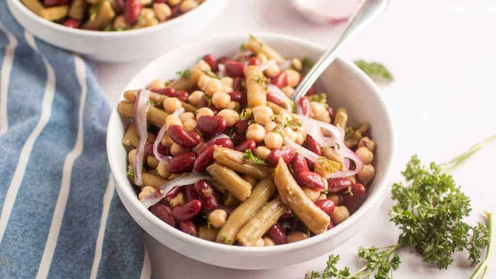 A bowl of three-bean salad with kidney beans, chickpeas, onions, and herbs sits on a table next to a blue striped napkin and fresh parsley.