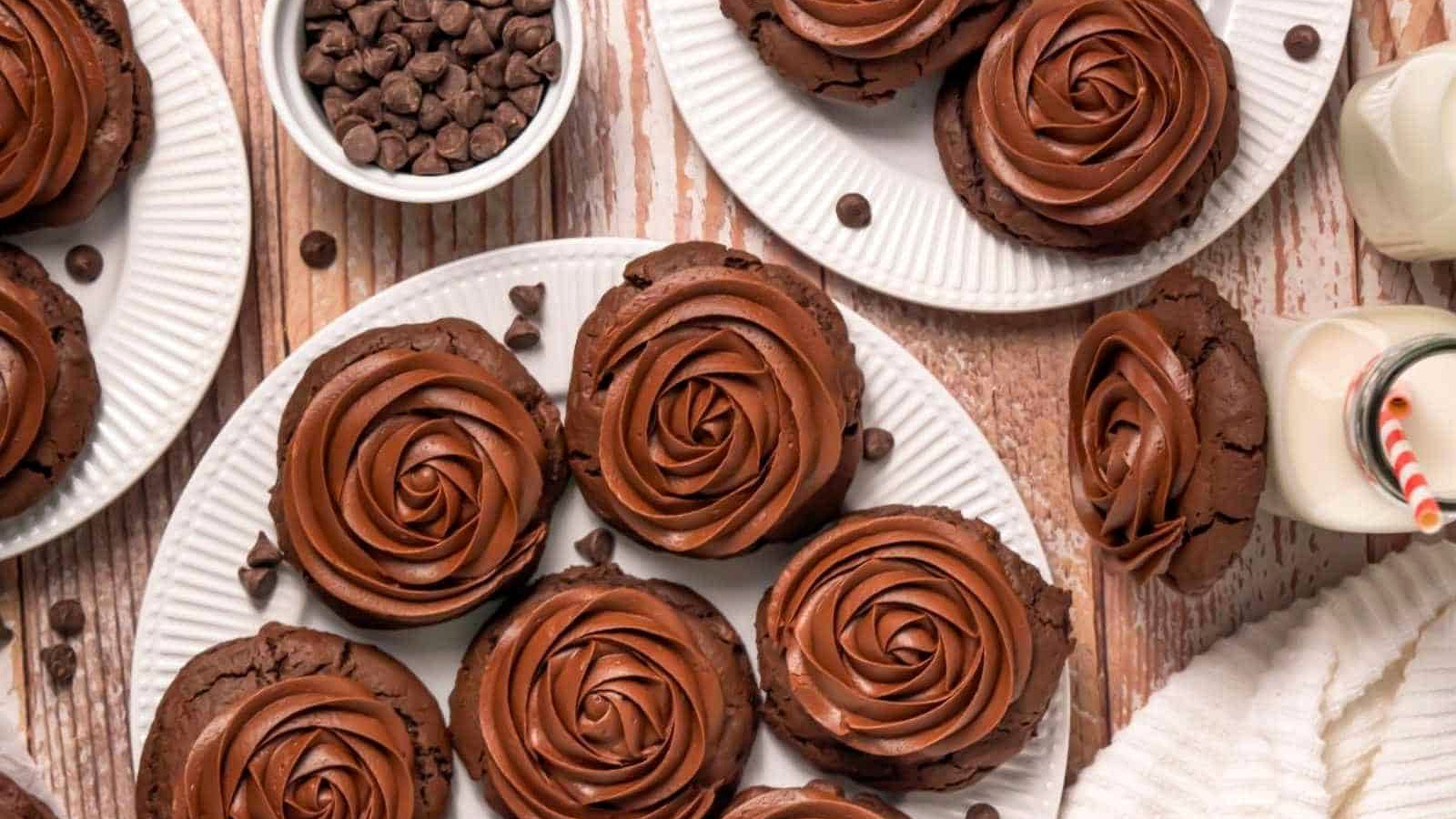 Plates of chocolate cookies with swirled chocolate frosting, a bowl of chocolate chips, and bottles of milk with striped straws on a wooden surface.