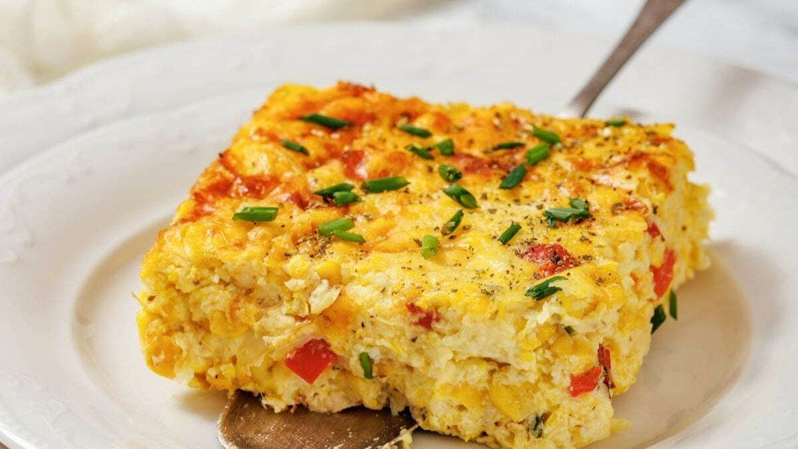 A square slice of corn pudding casserole with eggs, red bell pepper, and chives sits on a white plate with a serving spoon underneath.