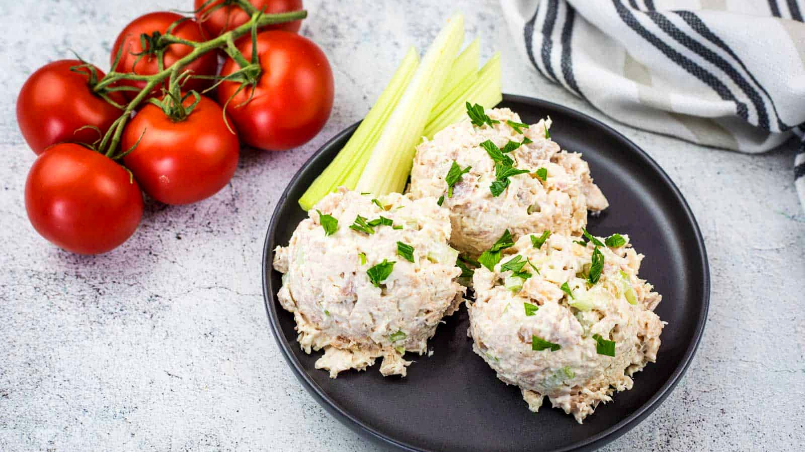A black plate with three scoops of Copycat Costco Chicken Salad garnished with parsley, celery sticks, and a bunch of tomatoes on the side. A striped towel is in the background.