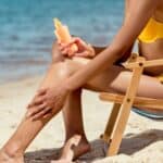A person in a yellow bikini sits on a beach chair by the sea, applying sunscreen to their leg.