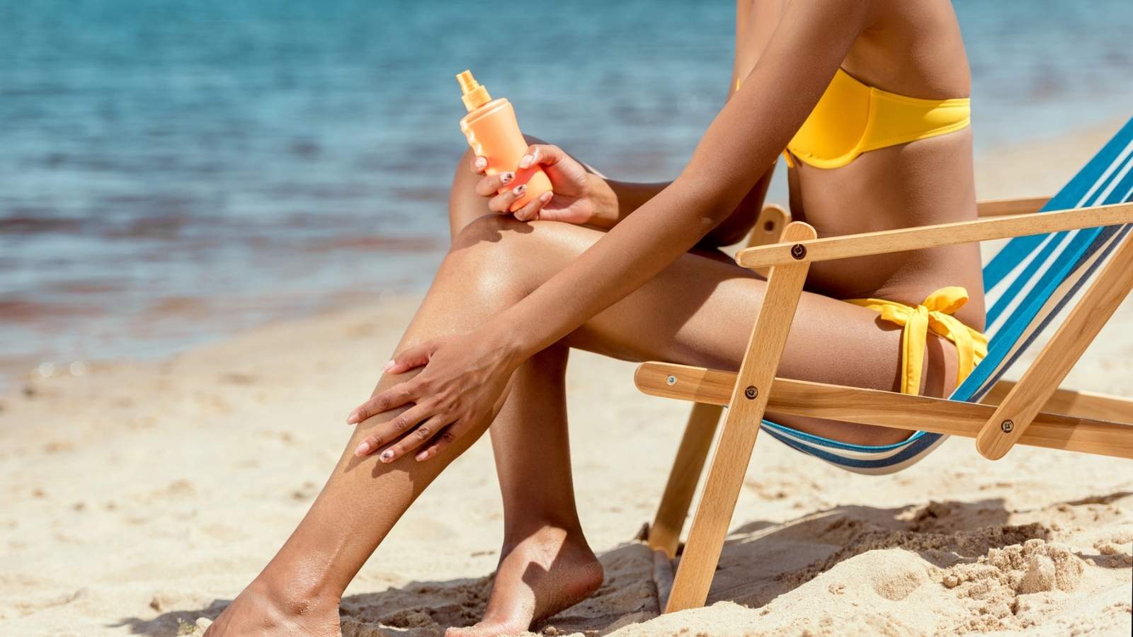 A person in a yellow bikini sits on a beach chair by the sea, applying sunscreen to their leg.