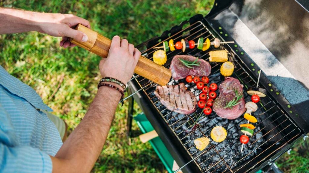 Person seasoning steaks and vegetables, including corn, tomatoes, and peppers, on an outdoor grill with an open lid. Grass is visible in the background.