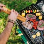 Person seasoning steaks and vegetables, including corn, tomatoes, and peppers, on an outdoor grill with an open lid. Grass is visible in the background.