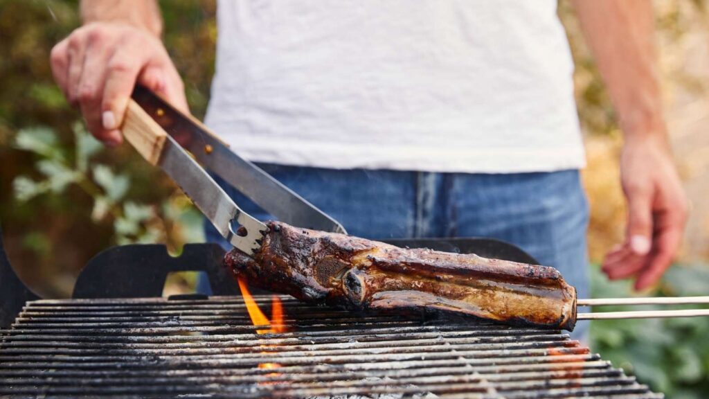 A person in a white shirt uses tongs to turn a large piece of meat on a grill over an open flame outdoors.