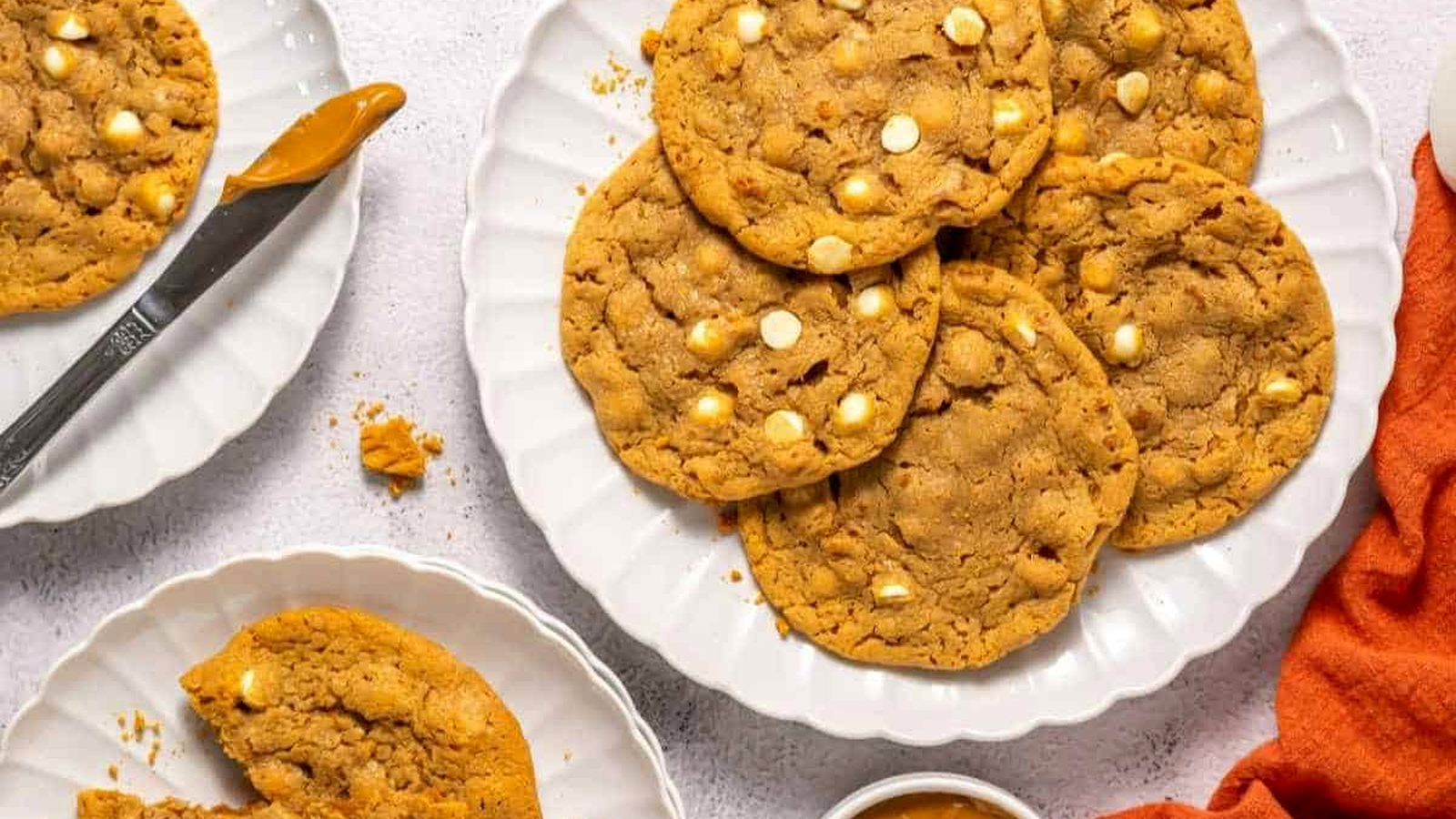 A plate of large peanut Copycat Crumbl Biscoff cookies with white chocolate chips, surrounded by a knife, a small bowl of peanut butter, and an orange napkin.