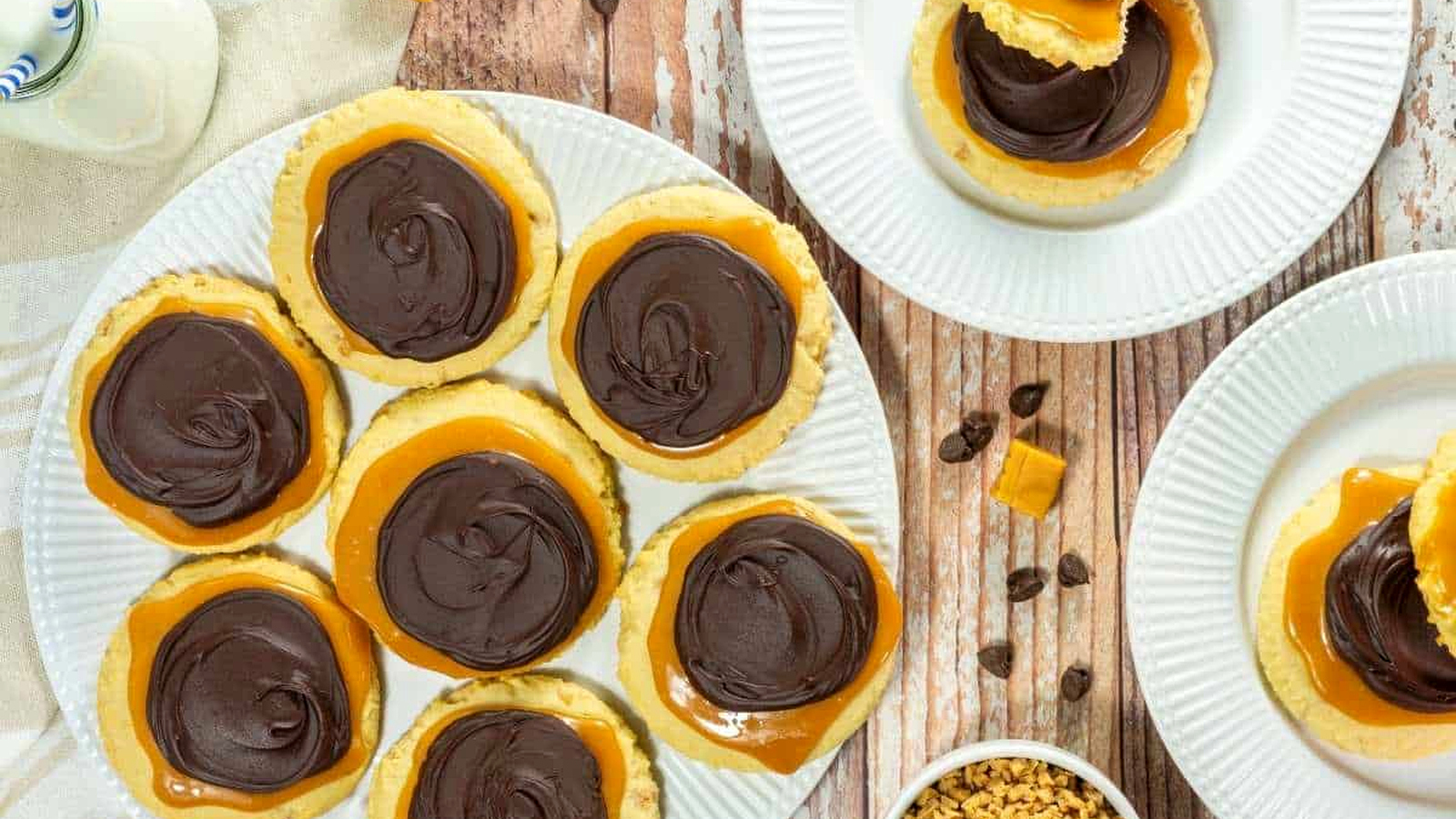 A plate of Copycat Crumbl Twix Cookies, with more cookies on separate plates, caramel pieces, and small chocolate chips nearby on a wooden table.