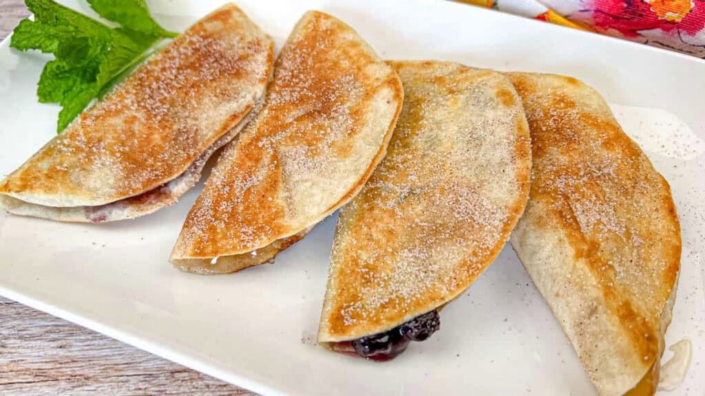 Four folded, lightly browned tortillas with a dusting of sugar are arranged on a white plate, garnished with a sprig of mint.