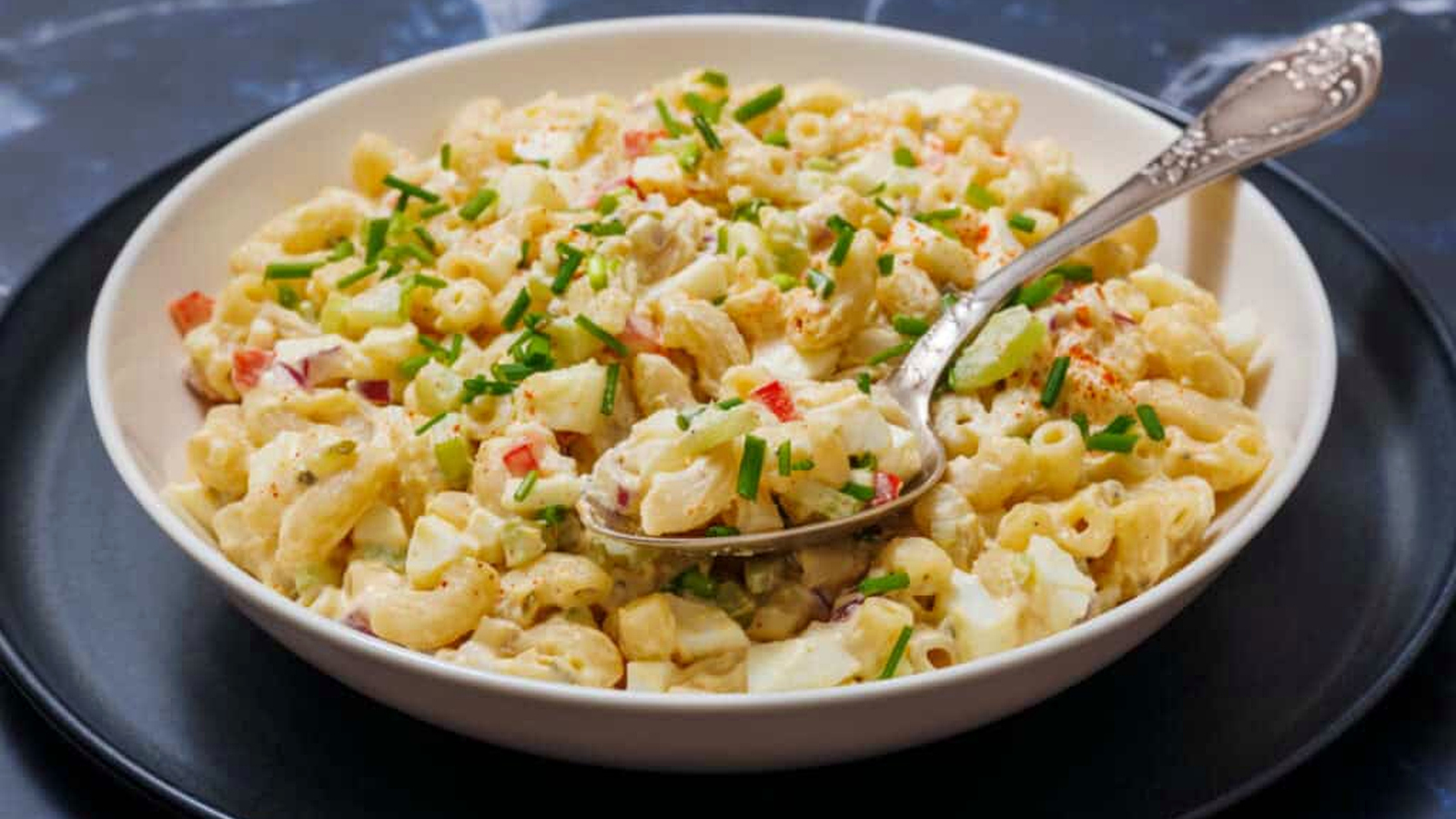 A bowl of Deviled Egg Pasta Salad with chopped vegetables and herbs, is set on a black plate, and a spoon rests in it.