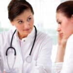 A doctor with a stethoscope consults with a concerned woman, discussing information on a clipboard in a bright medical office.