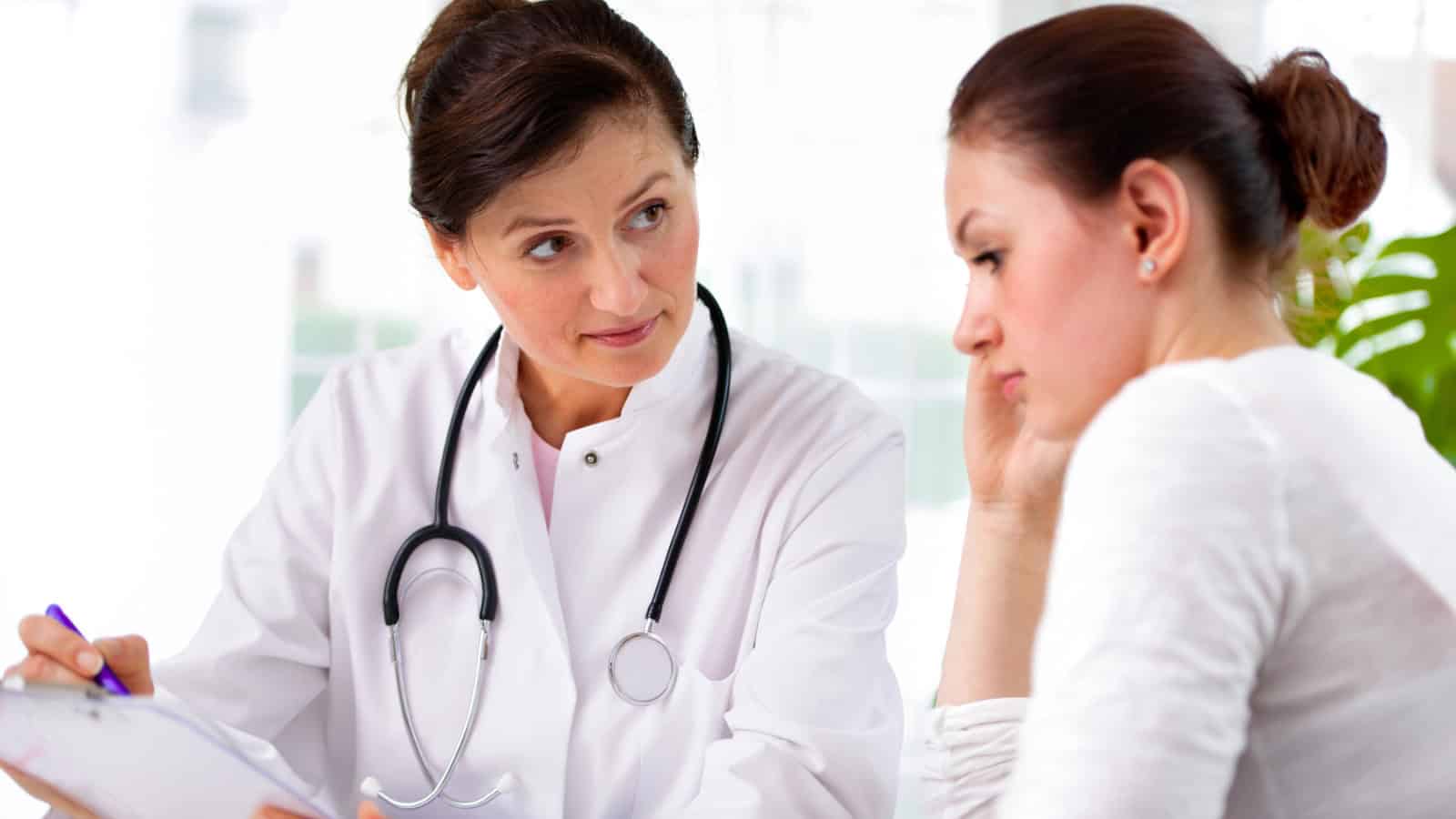 A doctor with a stethoscope consults with a concerned woman, discussing information on a clipboard in a bright medical office.