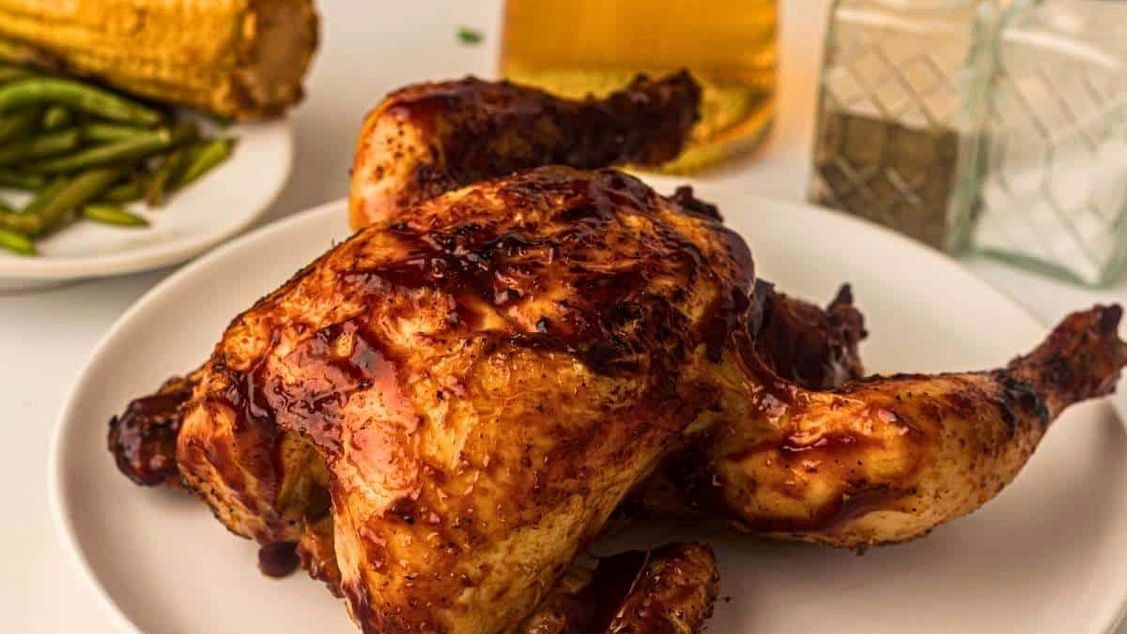 A roasted chicken with barbecue sauce on a white plate, with green beans, corn on the cob, a glass of beer, and salt and pepper shakers in the background.