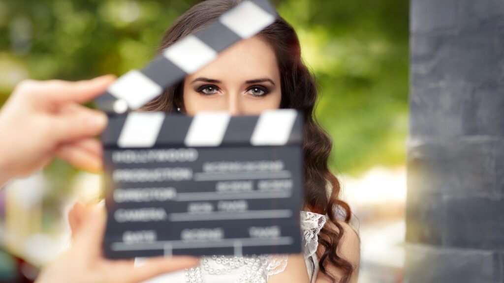 A person with long brown hair is partially obscured by a film clapperboard being held in front of their face outdoors.