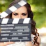 A person with long brown hair is partially obscured by a film clapperboard being held in front of their face outdoors.