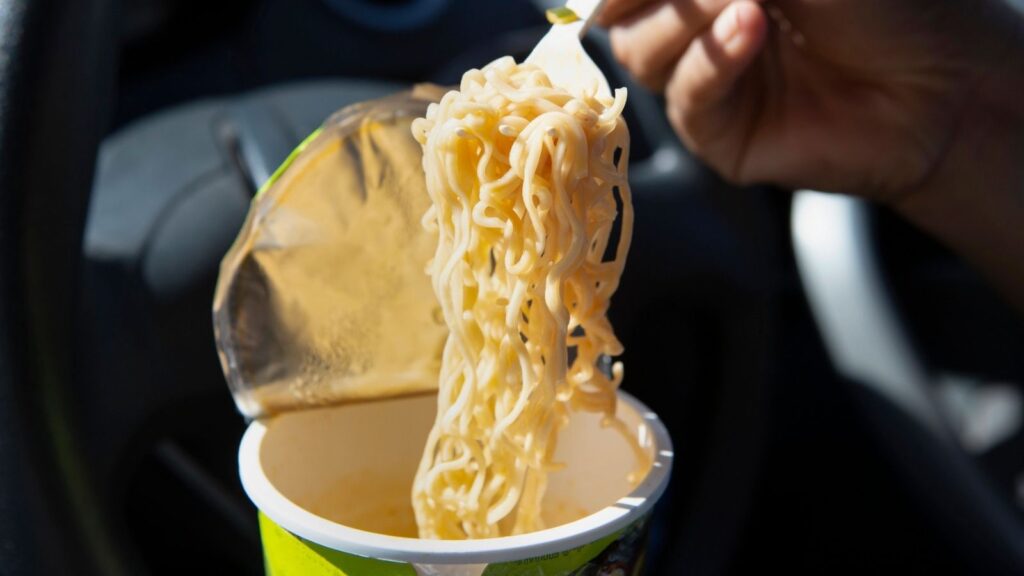 A hand holds a fork lifting cooked instant noodles from a cup with a partially opened foil lid, inside a car.