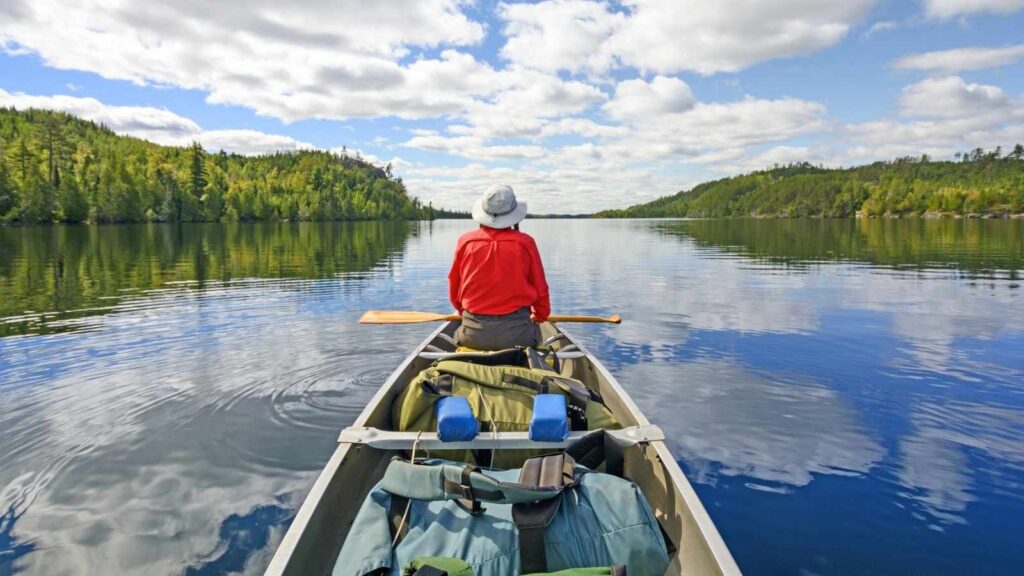A person wearing a red jacket and hat paddles a canoe on a calm lake surrounded by forest under a partly cloudy sky.