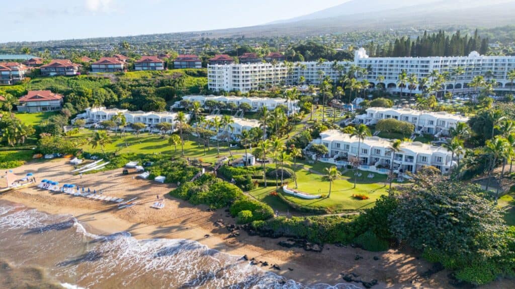 Aerial view of a beachfront resort with white buildings, green lawns, palm trees, and beach chairs facing the ocean, with mountains and houses in the background.