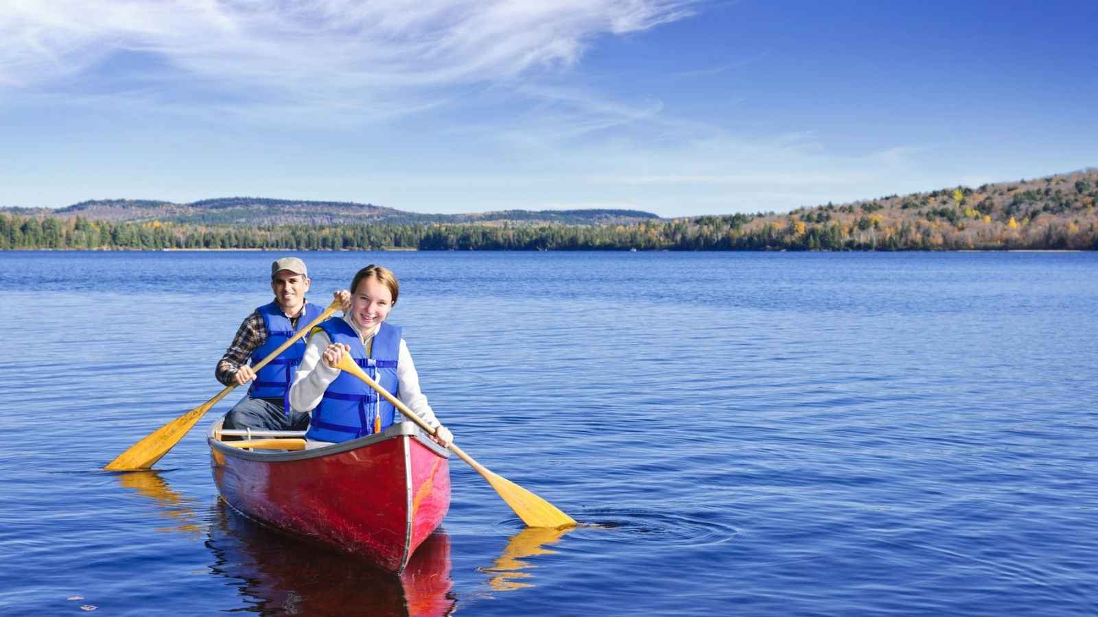 Two people paddle a red canoe on a calm lake surrounded by forested hills under a clear blue sky.