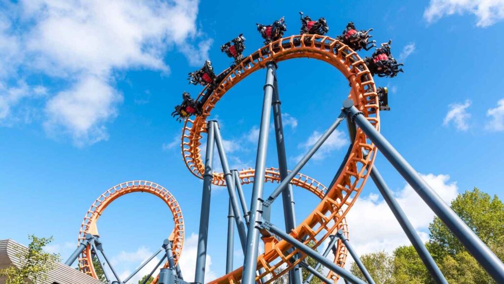 People ride a looping orange and blue roller coaster under a bright blue sky with scattered clouds.