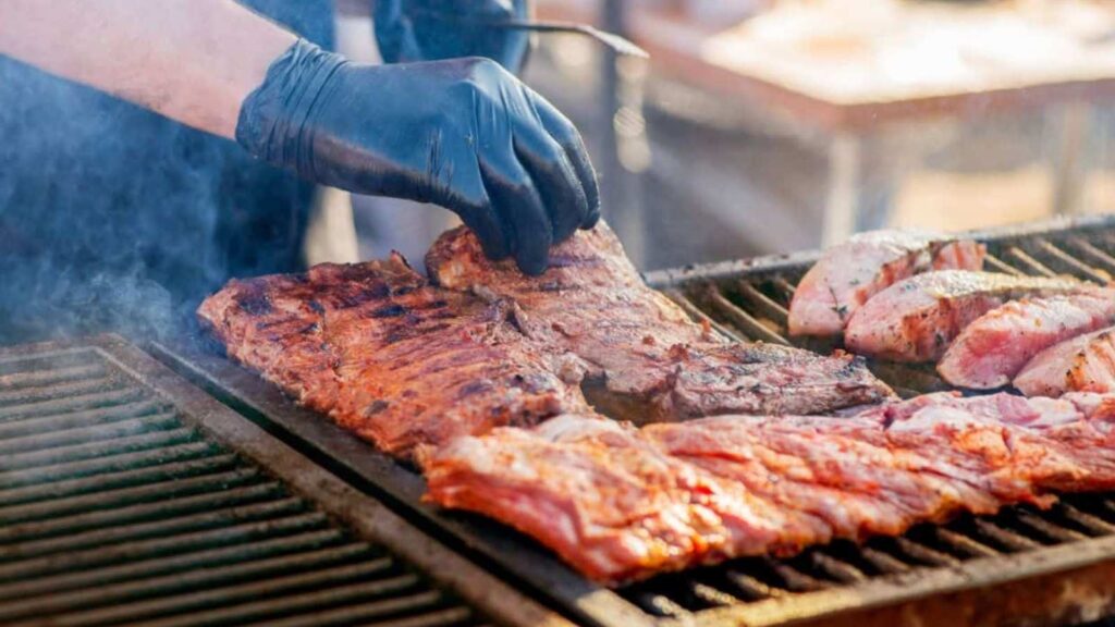 Person wearing a black glove grills racks of ribs and seasoned chicken pieces on an outdoor barbecue with visible smoke, showcasing expert grilling tips for foods.