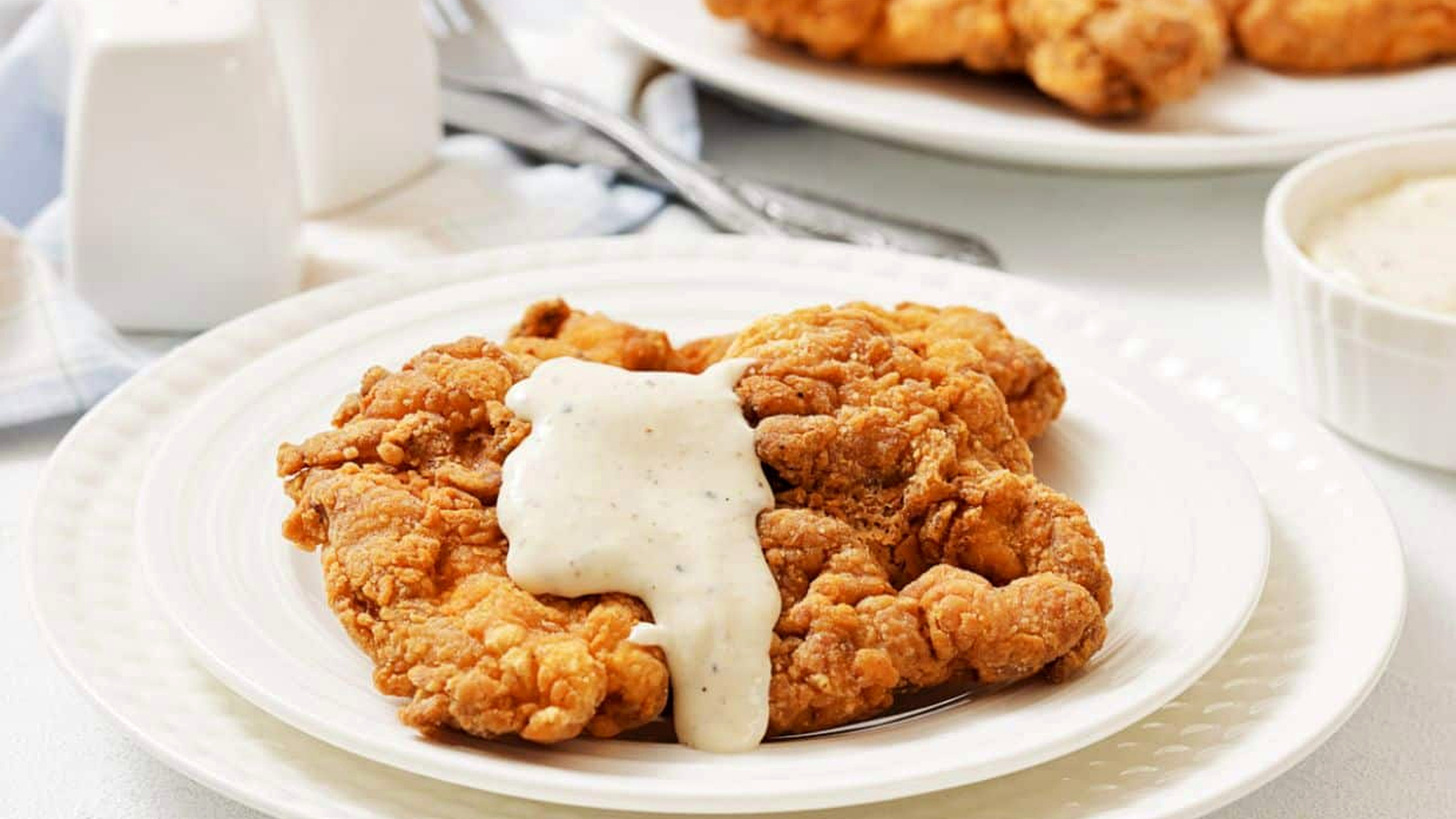 A plate of fried chicken steak topped with white gravy, with more fried pieces and a bowl of gravy in the background.