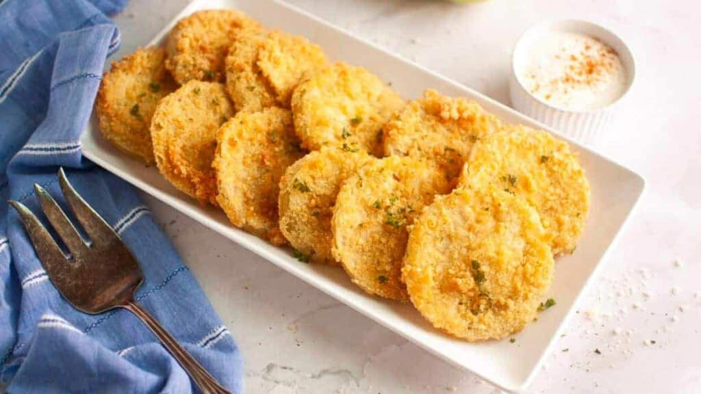 A rectangular white plate of breaded and fried green tomato slices garnished with herbs, next to a cup of dipping sauce and a fork on a blue cloth.