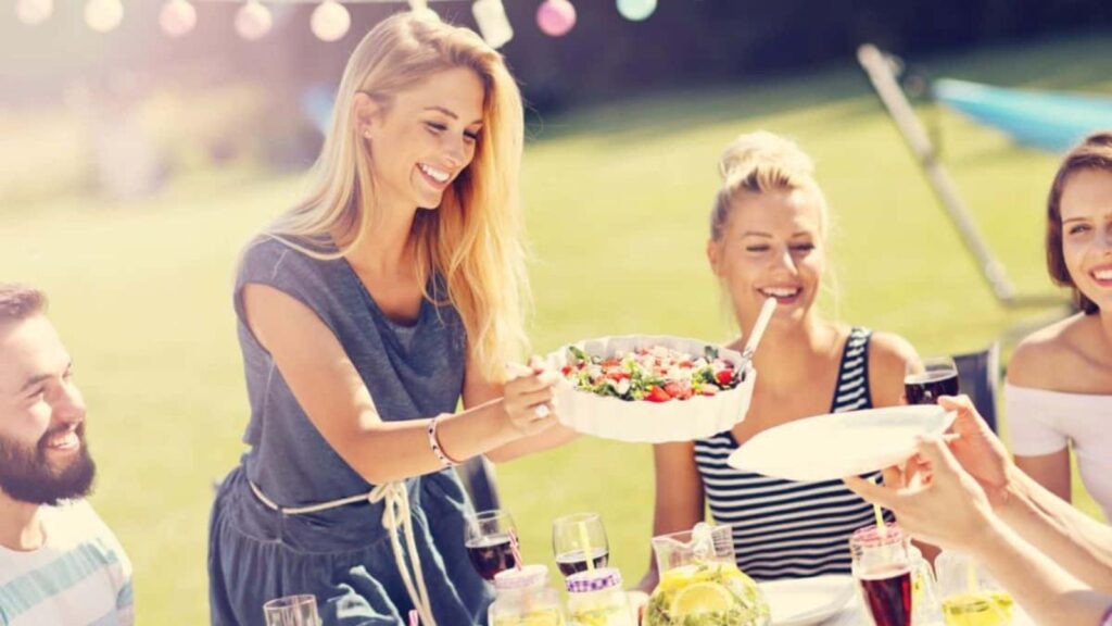 A woman serves salad to people at an outdoor table, where several individuals are gathered, smiling and enjoying dining out together in the warm sunlight.