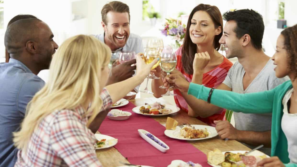 Six people sit around a table sharing a meal and clinking glasses, smiling and engaging in conversation in a bright, casual setting.