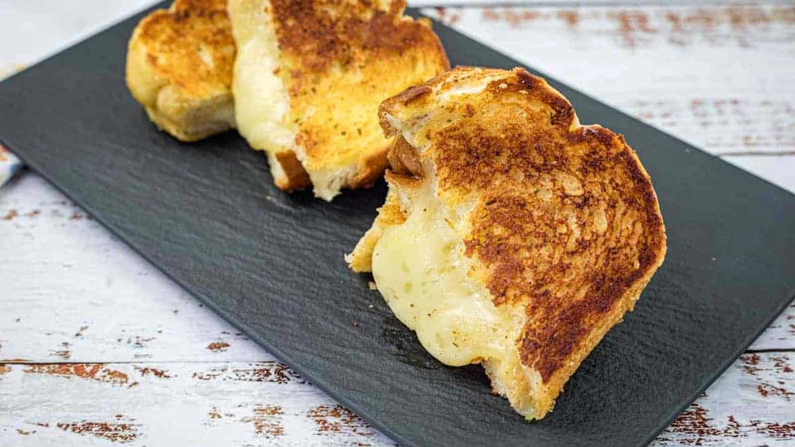 Three garlic bread grilled cheese on a black slate plate on a white wooden surface.