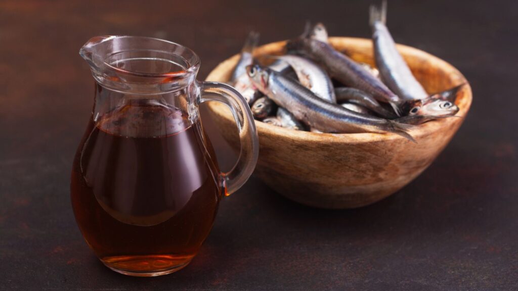 A glass jug filled with dark fish sauce, reminiscent of ancient Roman garum, sits next to a wooden bowl containing several whole raw fish on a dark surface.