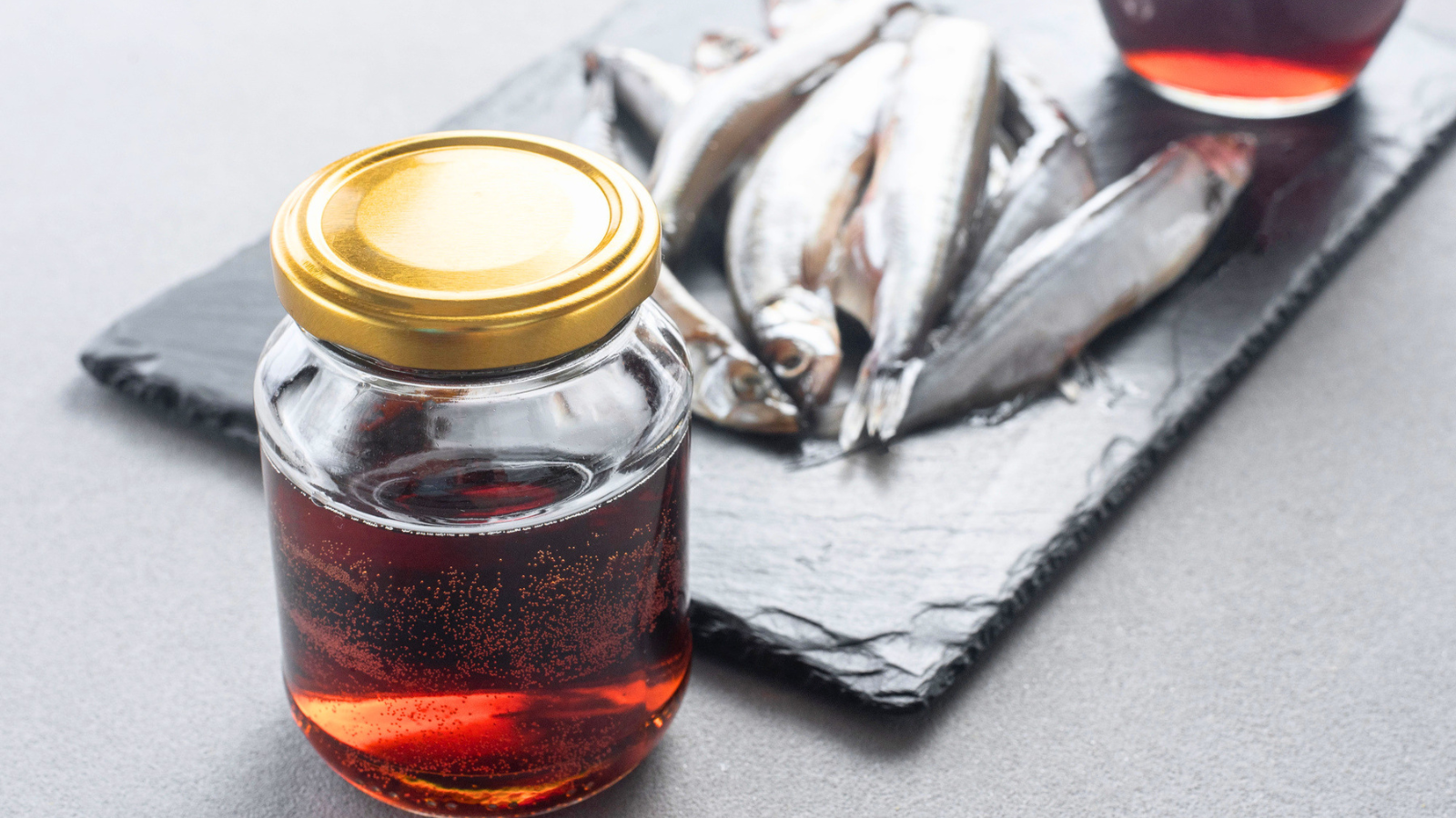 A glass jar filled with amber-colored fish sauce, reminiscent of Ancient Roman garum, sits on a gray surface next to a slate platter with several small raw fish.