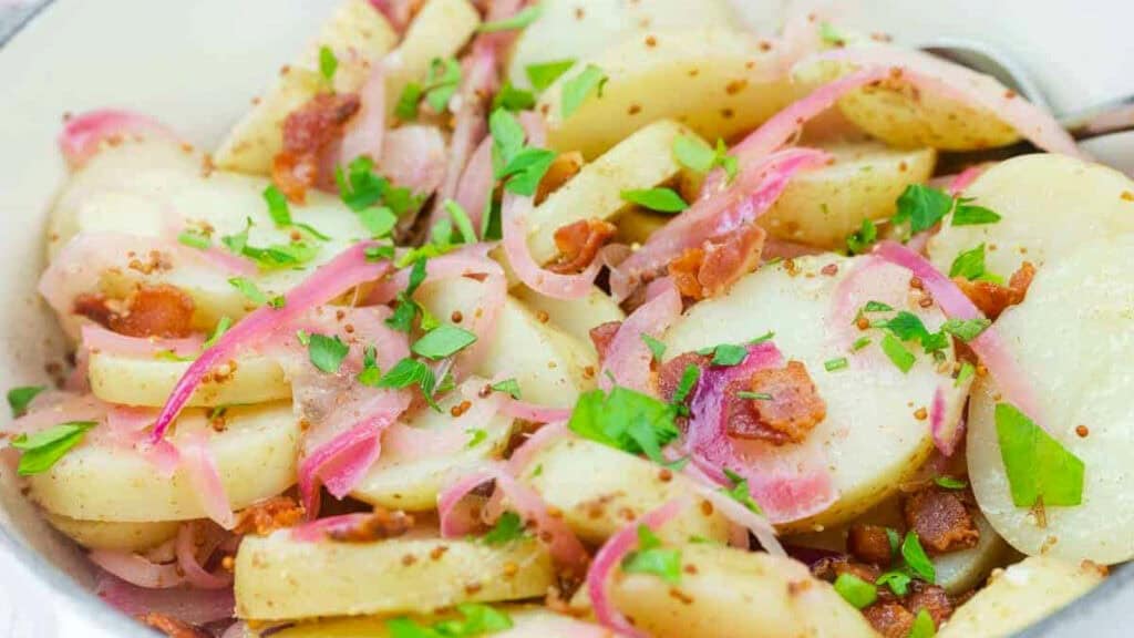 Sliced potatoes with red onions, bacon pieces, and chopped parsley, mixed in a light dressing and served in a bowl.