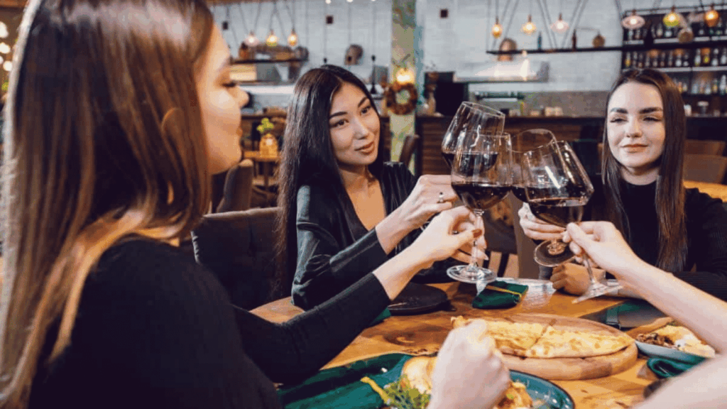 Four women sit at a restaurant table, raising glasses of red wine in a toast. Plates of food are in front of them, and the setting appears warm and casual.