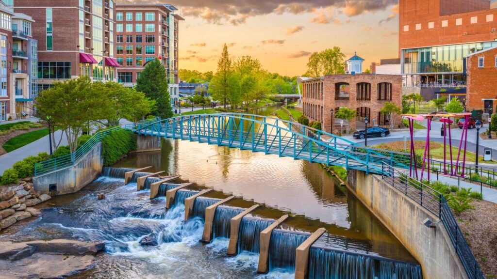 A footbridge crosses over a small river with cascading water in a city park, where modern and historic buildings at sunset reflect the charm of the Carolinas—a perfect scene after watching Top Chef Carolinas.