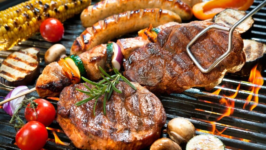 Assorted grilled meats, sausages, vegetables, and corn on a barbecue grill with flames underneath.