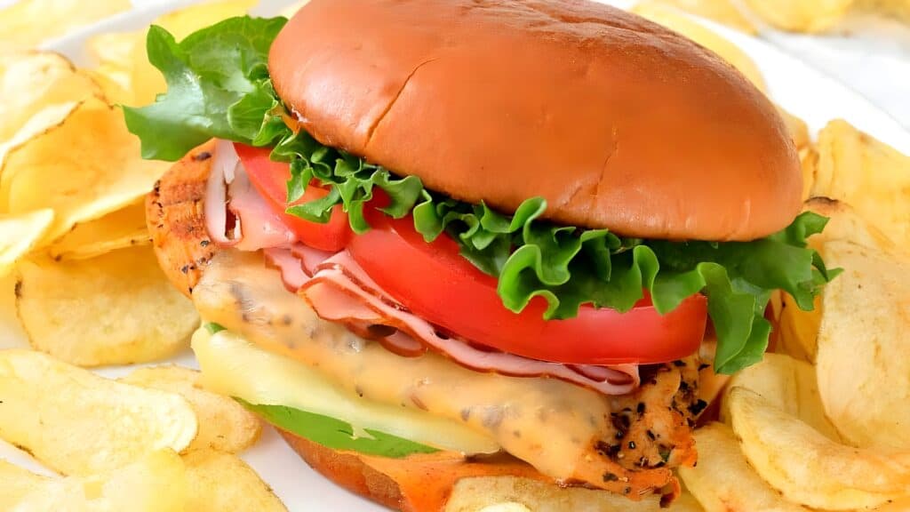 A grilled chicken sandwich with lettuce, tomato, cheese, and sliced ham on a bun, served with potato chips.