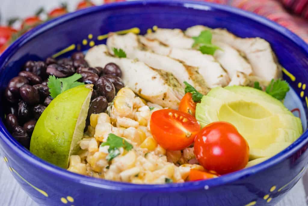 A blue bowl filled with sliced grilled chicken, black beans, avocado, lime, corn salad, and cherry tomatoes, garnished with cilantro.
