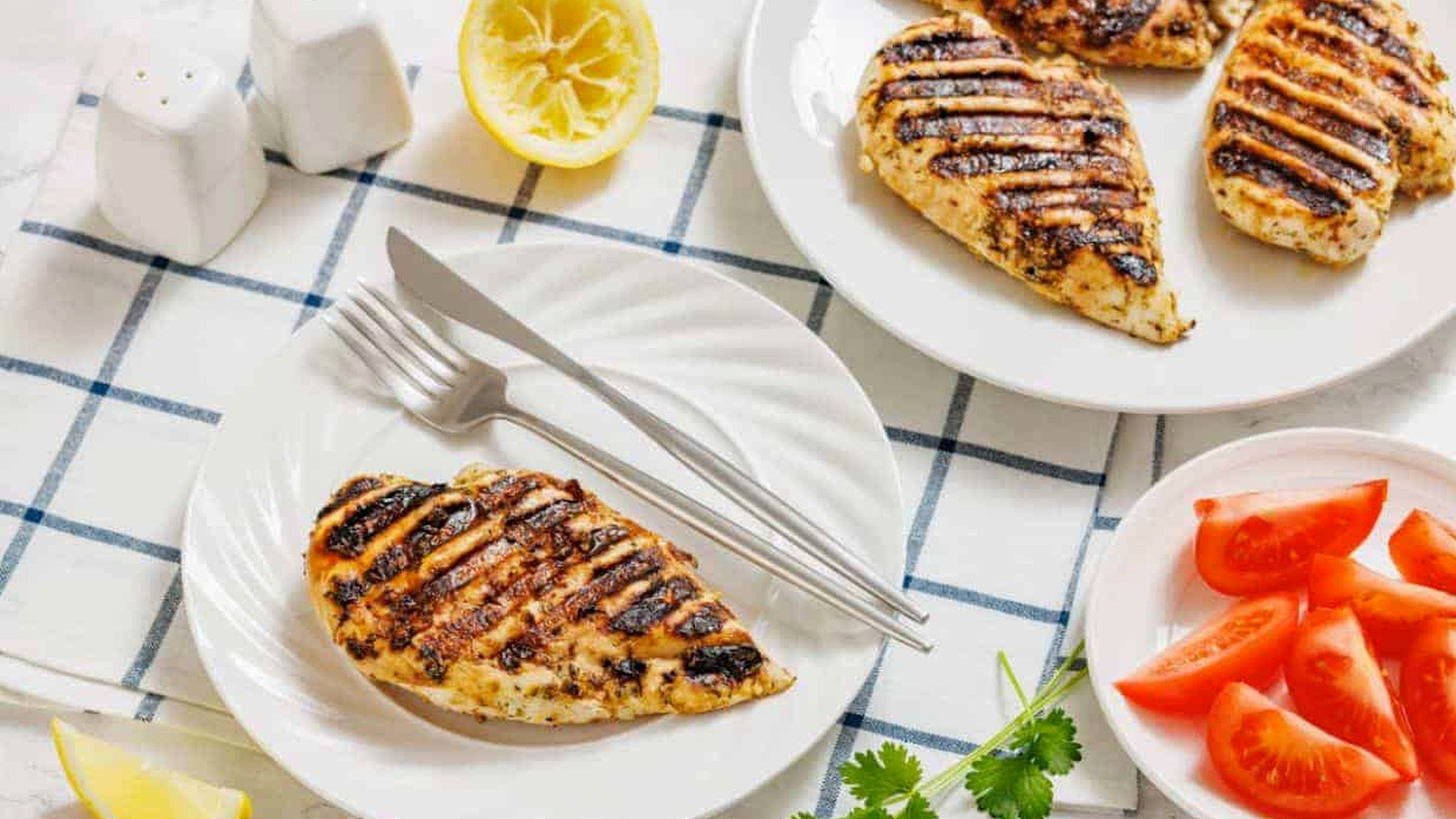 Grilled chicken breast on a white plate with fork and knife, sliced tomatoes, lemon halves, cilantro, and salt and pepper shakers on a checkered cloth.