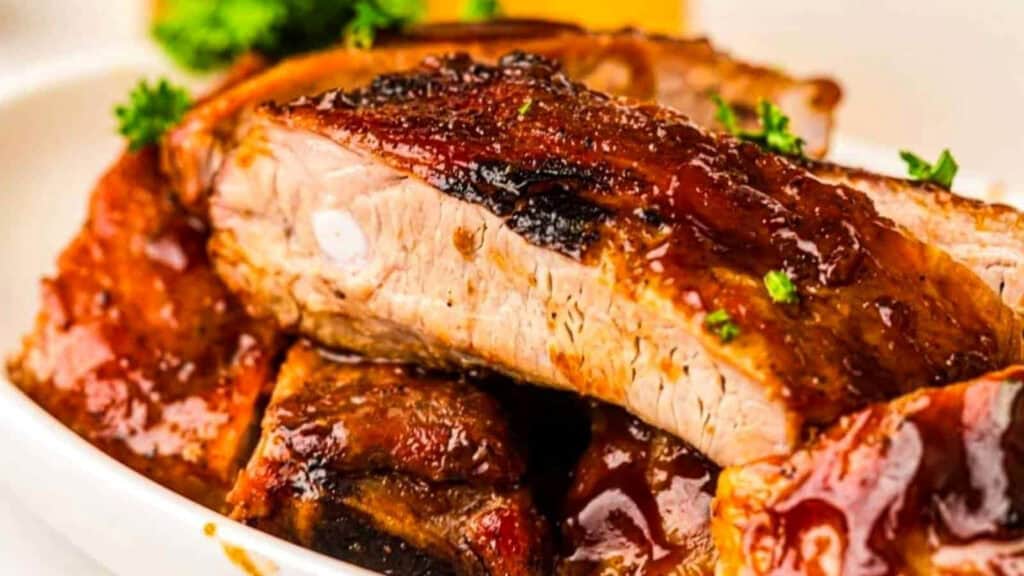 Close-up of barbecued pork ribs with a caramelized glaze, garnished with small sprigs of parsley on a white plate.